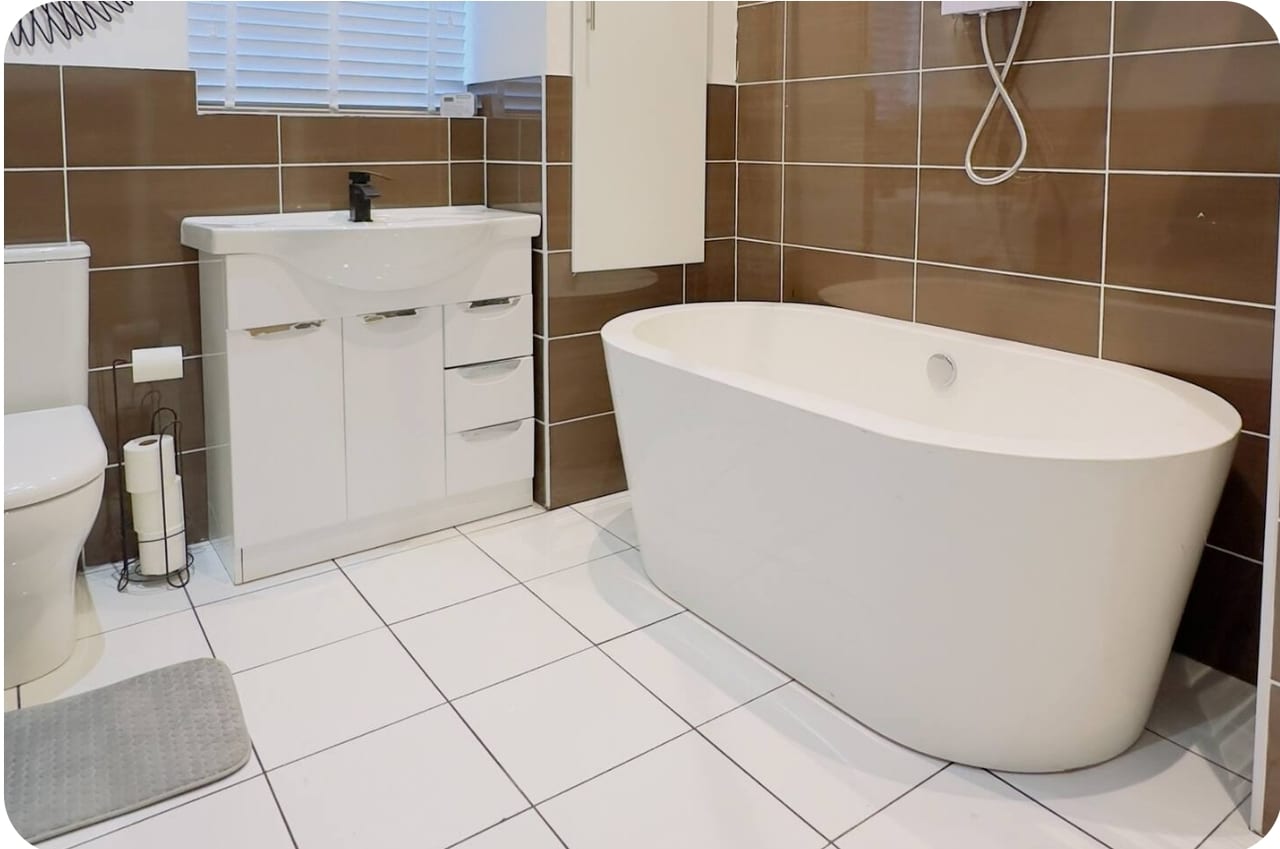 White freestanding bath beside a basin cabinet, with brown wall tiles and a tiled floor.