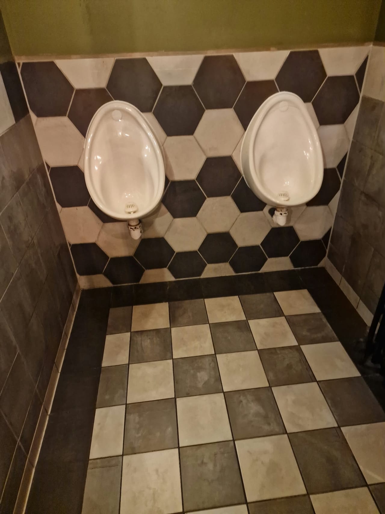 Two wall-mounted white urinals against a geometric tiled wall above a chequered floor.