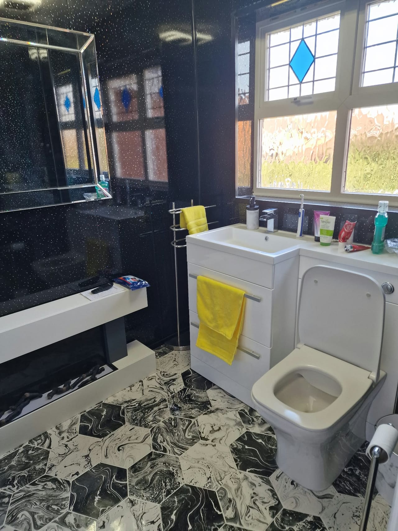 White bath and toilet beneath a decorative window, with dark walls and a patterned floor.