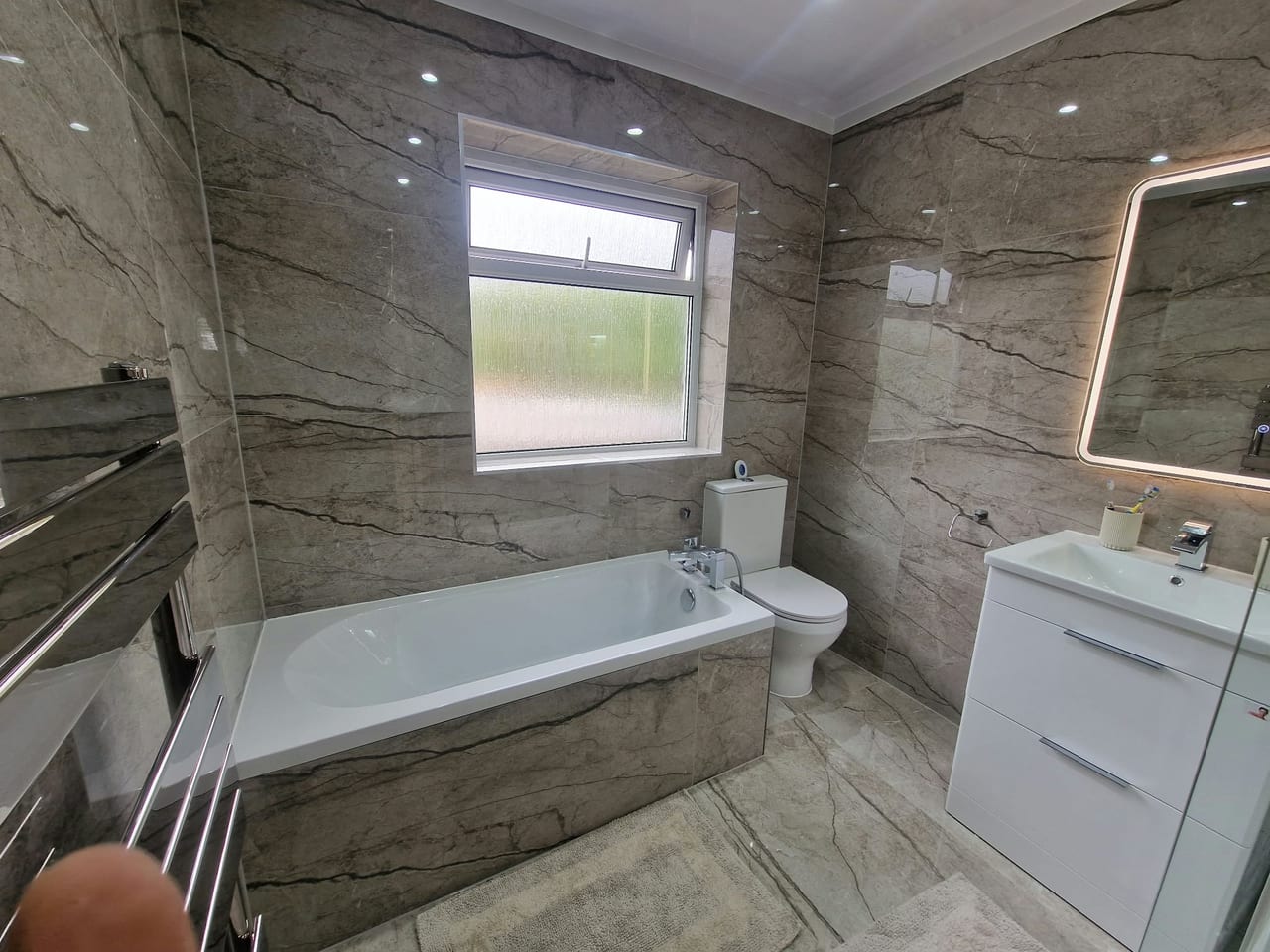 White bath beneath a window beside a basin cabinet, surrounded by grey marble-effect tiles.