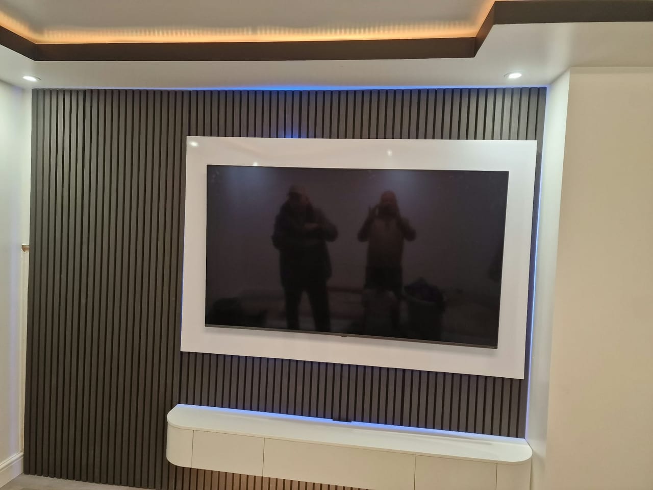 Wall-mounted television and white radiator on a dark panelled feature wall with inset lighting.