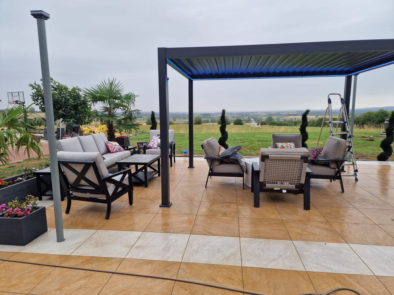 Paved terrace with outdoor seating beneath a dark pergola overlooking open countryside.