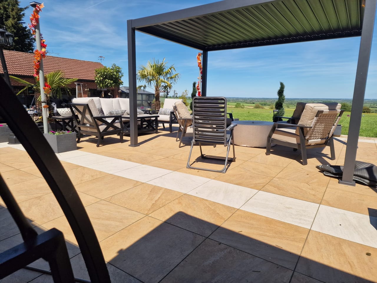 Paved terrace with outdoor seating beneath a dark pergola and views over open countryside.