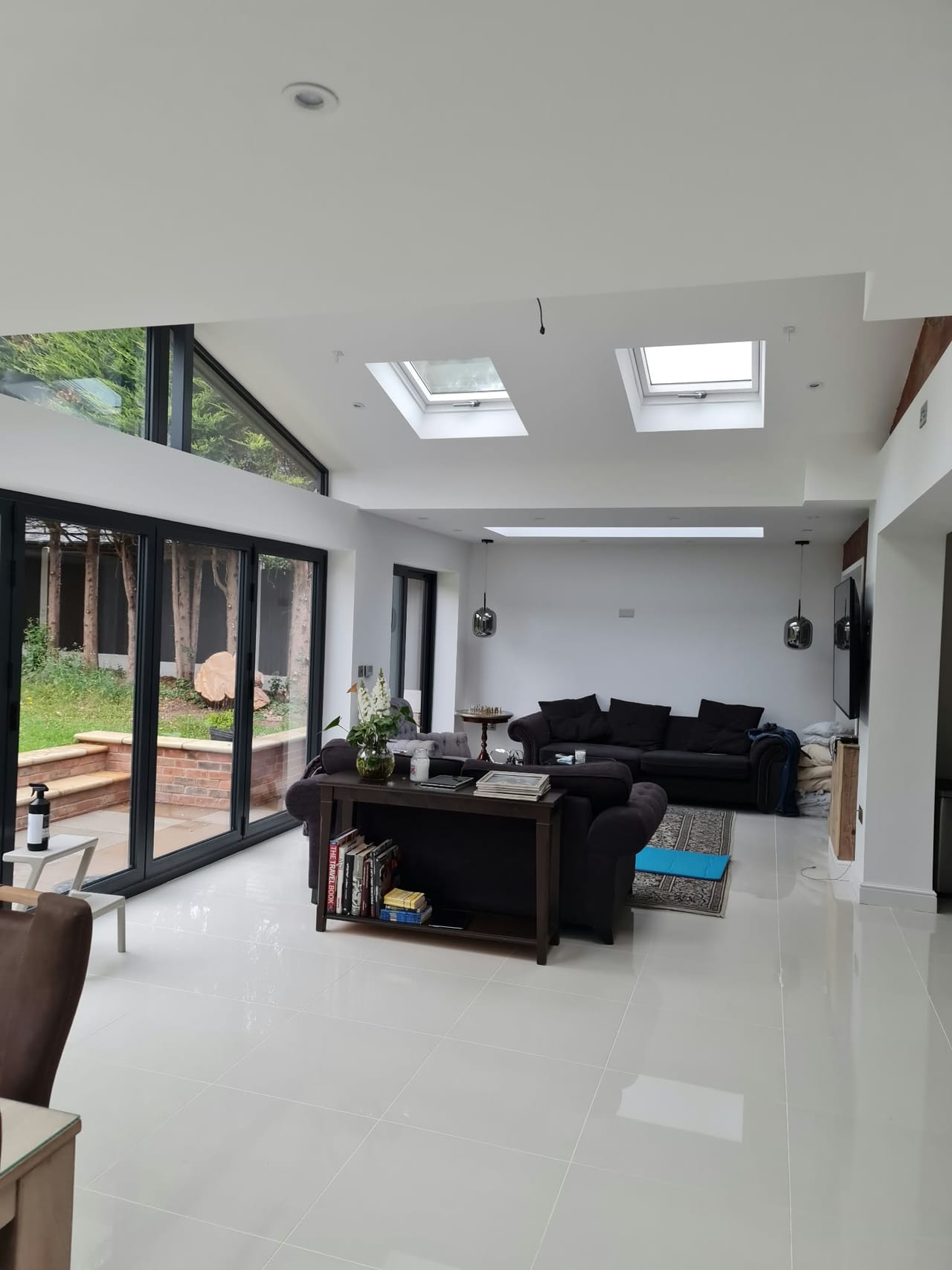Open-plan living space with rooflights, large glass doors, pale floor tiles and dark sofas.